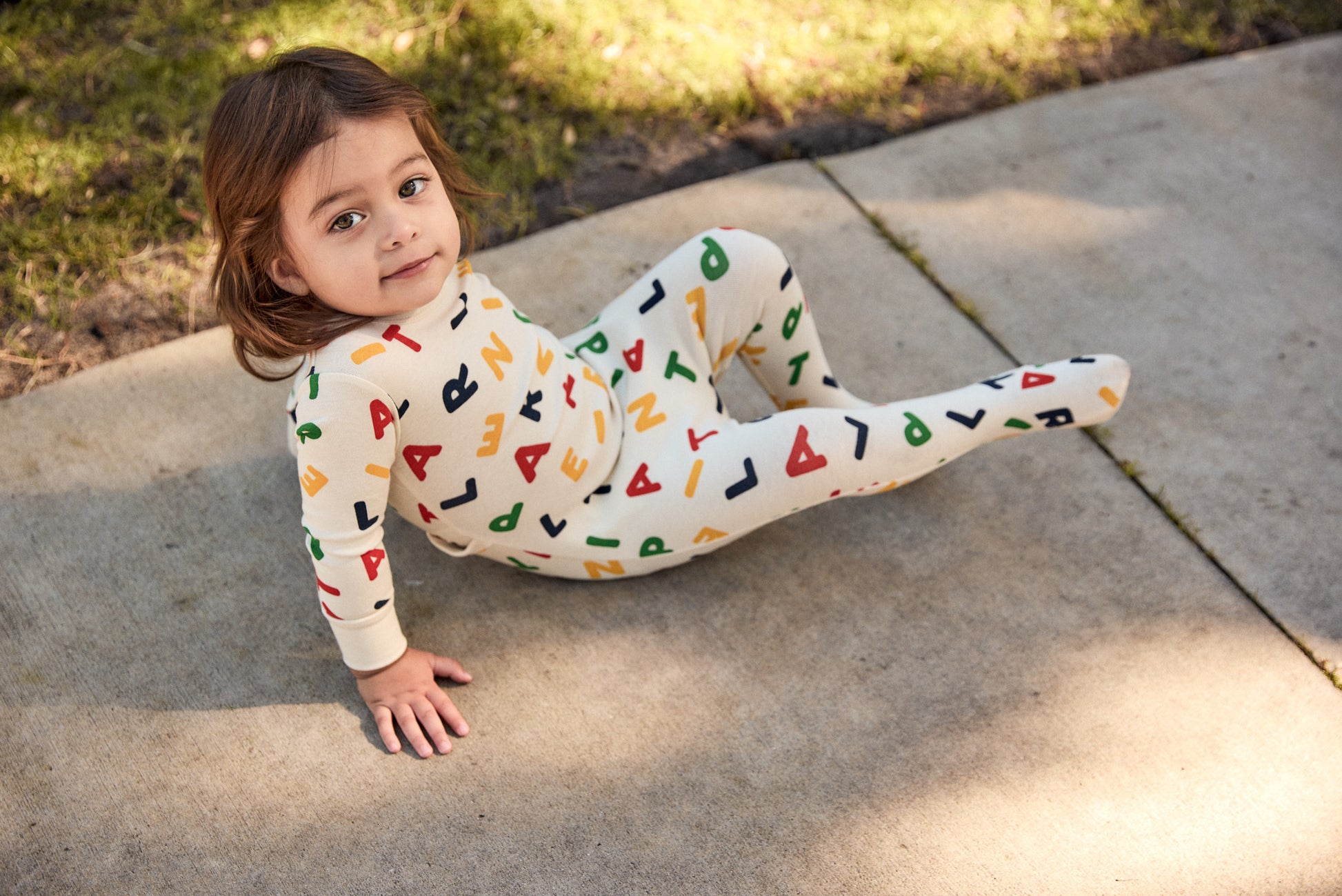 Child wearing a colorful onesie with letter patterns on a concrete surface.