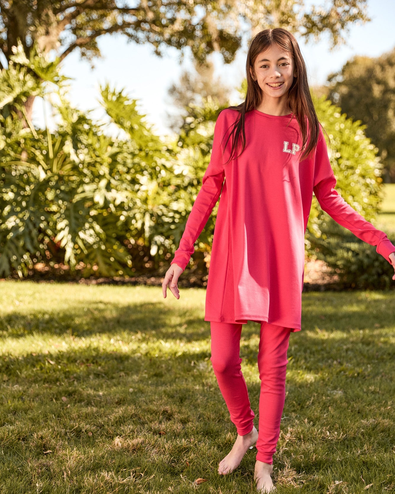 Teen wearing a pink outfit with 'L.P.' on it, standing in a grassy area with trees.