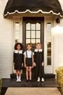 Three children standing on a porch with a black awning and door.