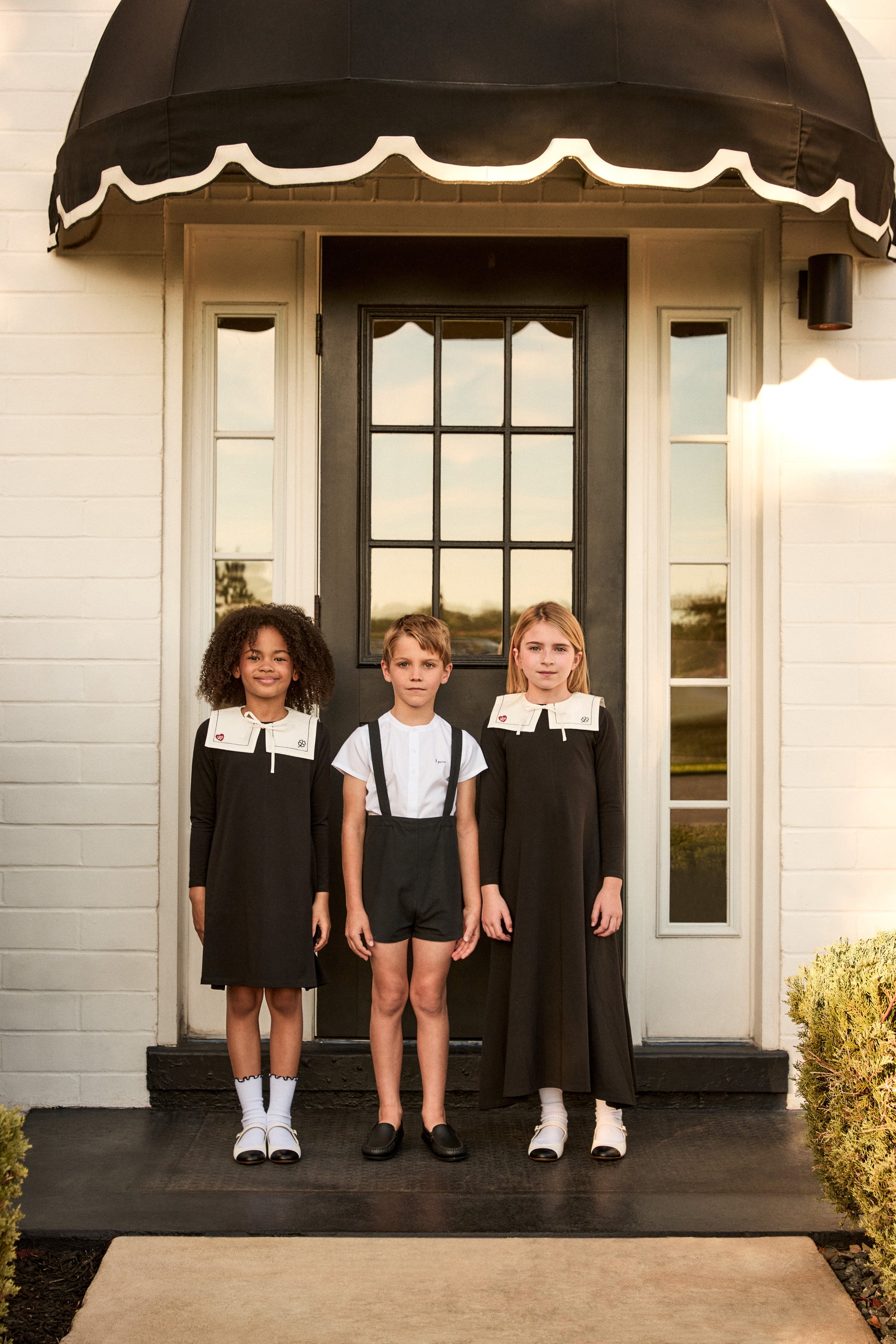 Three children standing on a porch with a black awning and door.