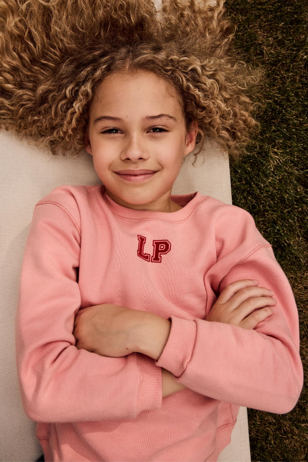 Child wearing a pink sweatshirt with 'LP' logo, lying on grass
