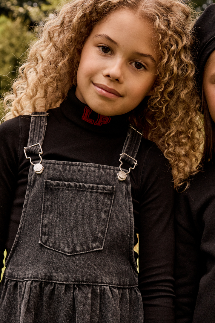 Young girl wearing a denim dress with suspenders, standing outdoors.