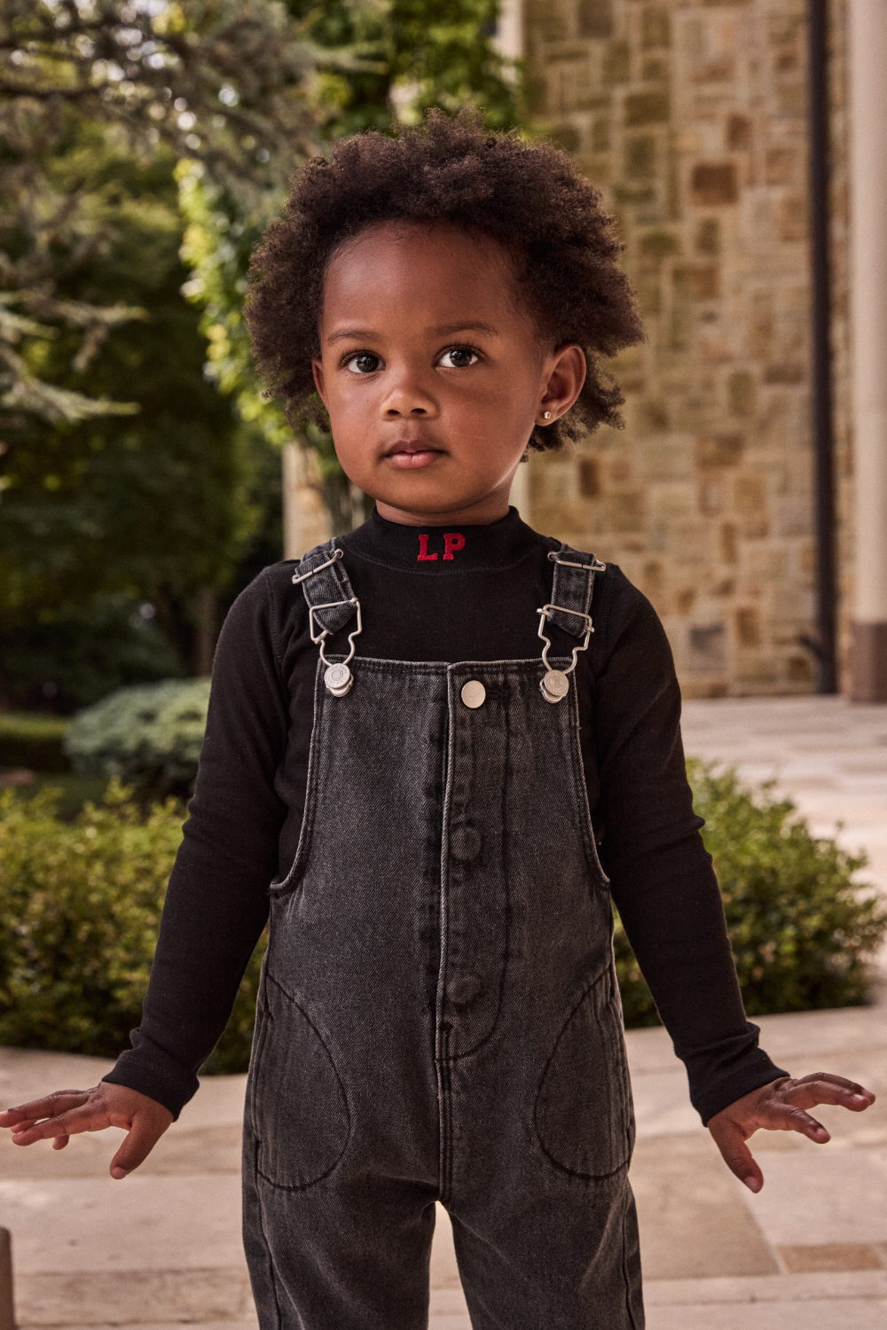 Child wearing a black turtleneck with 'LP' logo and gray overalls outdoors.