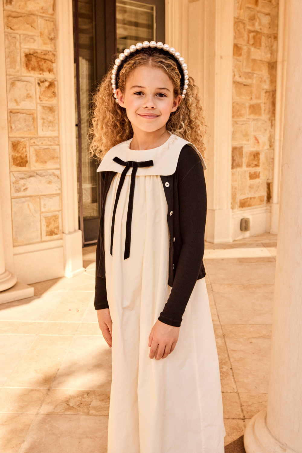 Young girl in a white dress with black accents standing in front of a stone building.