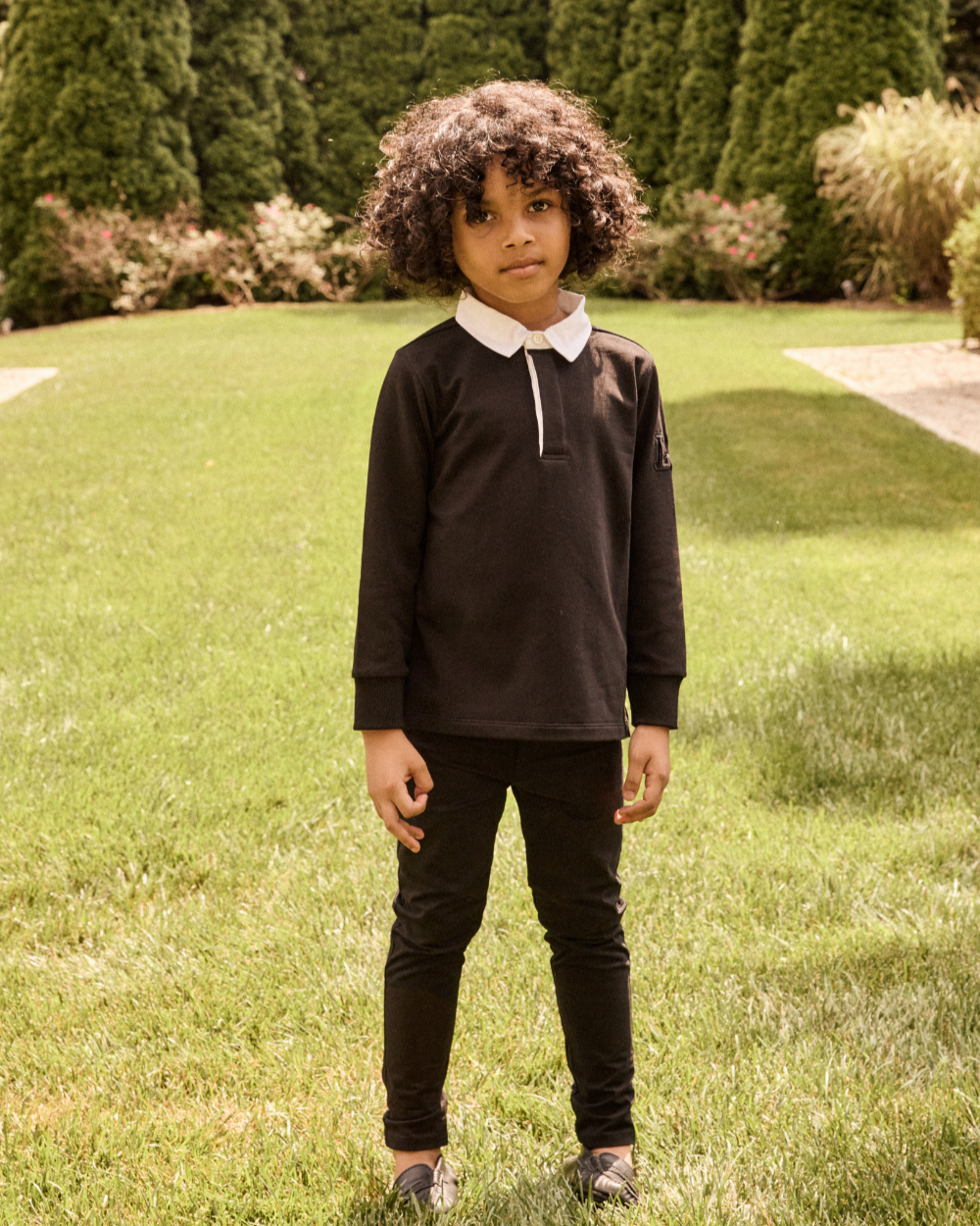 Child wearing a black long-sleeve shirt with a white collar in a grassy area with trees.
