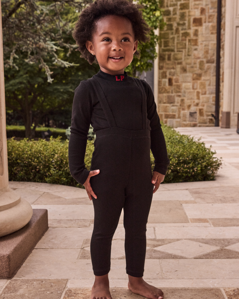 Child wearing a black outfit standing outdoors on a stone patio with greenery and a brick wall in the background.