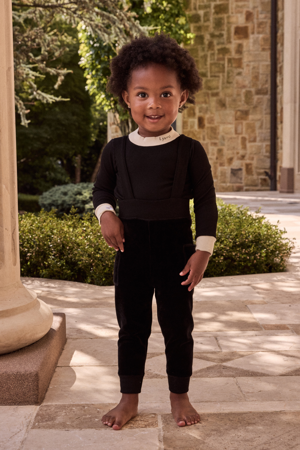 Child in a black outfit standing outdoors on a stone path with greenery and a stone building in the background.