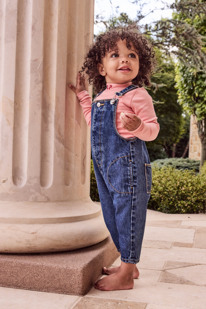 Child wearing denim overalls and a pink shirt standing outdoors near classical columns..