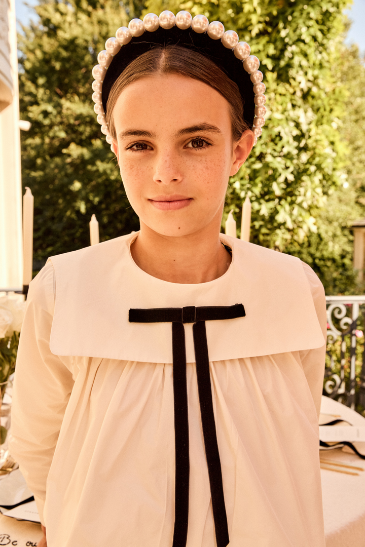 Young girl wearing a white dress with a black ribbon and a decorative headband outdoors.