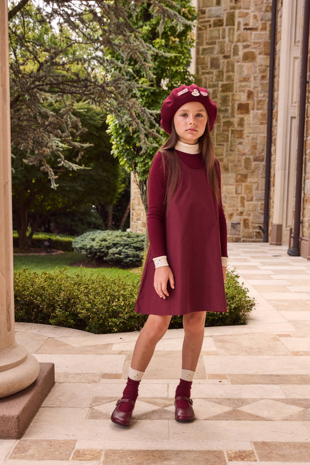 Young girl in a burgundy dress and hat standing outdoors on a stone path.