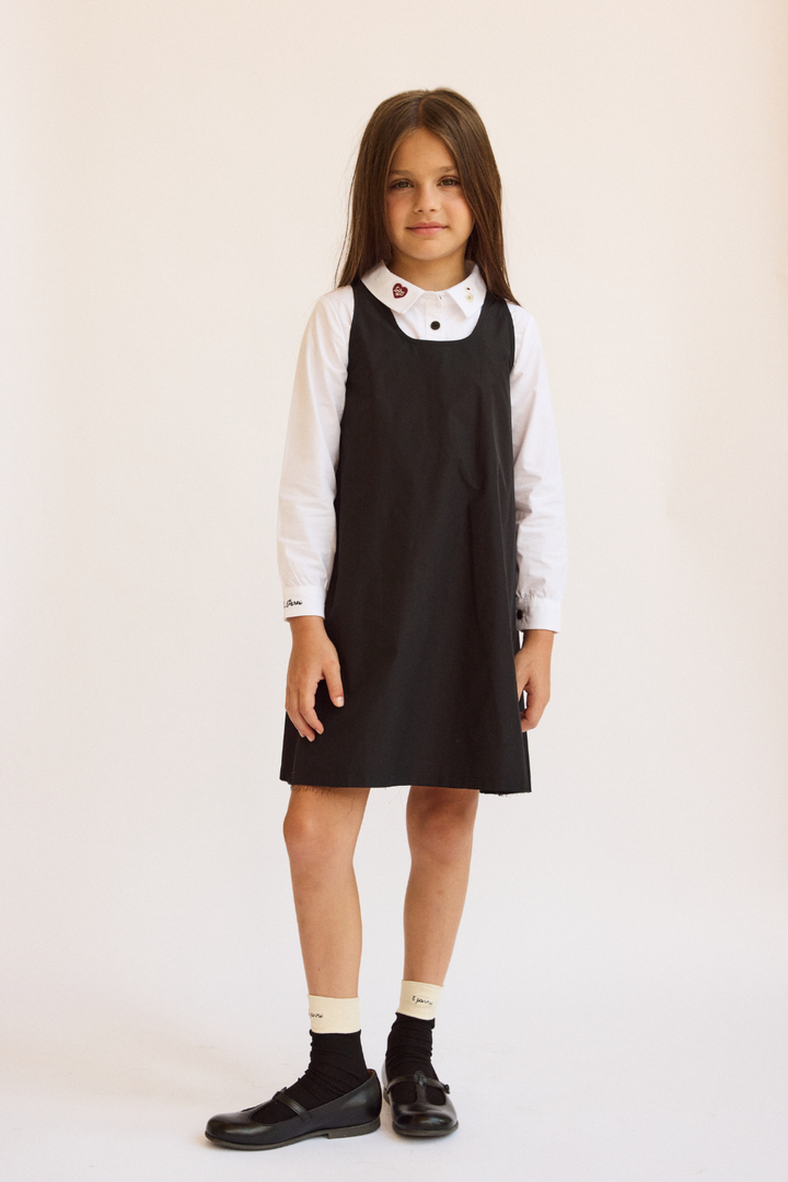 Young girl wearing a black pinafore dress over a white shirt on a plain background