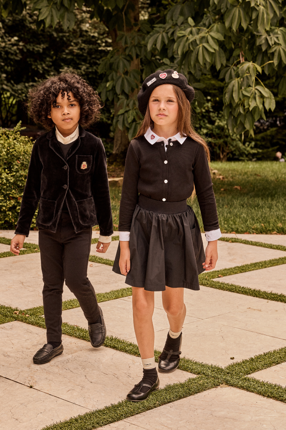 Two children in formal attire standing on a stone path with greenery in the background