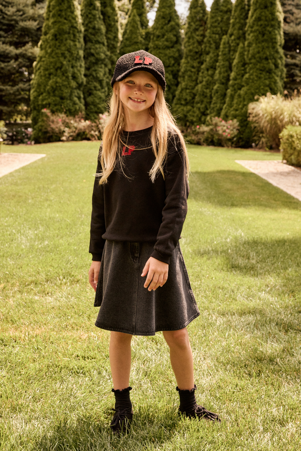 Young girl in a black outfit with a cap standing on grass.