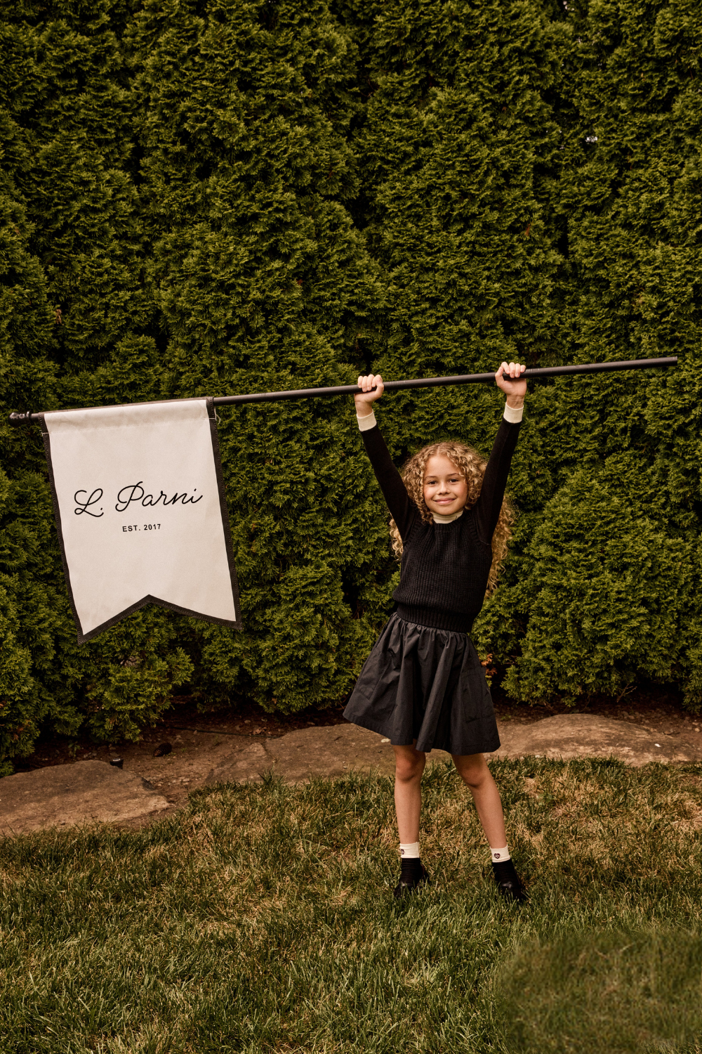 Young girl holding a banner with 'L' Parini' branding against a green hedge.