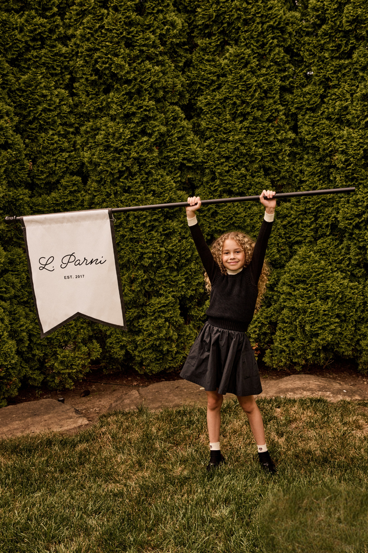 Young girl holding a banner with 'L' Parini' branding against a green hedge.