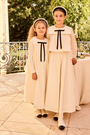 Two children in matching white dresses with black bows standing outdoors.