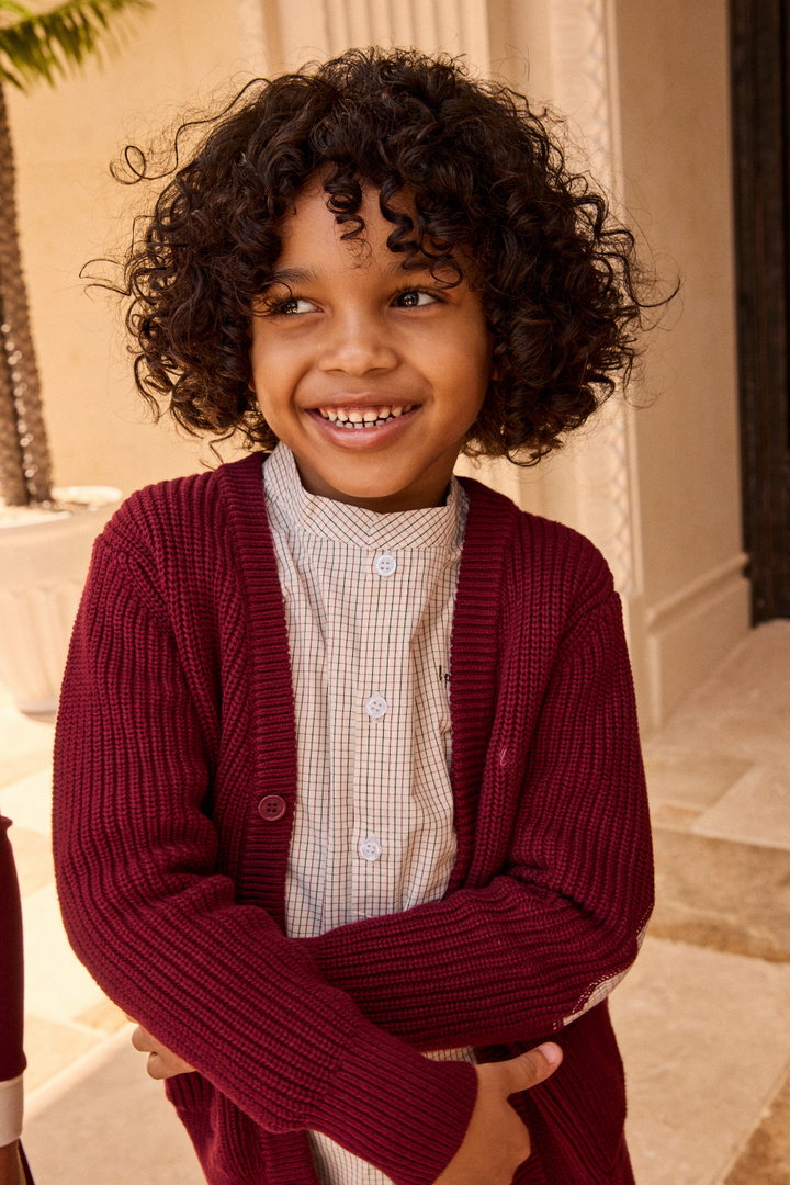 Child wearing a maroon cardigan and white shirt with a checkered pattern, standing outdoors.