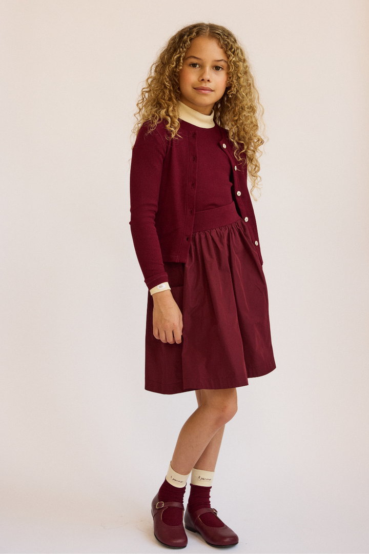 Young girl wearing a burgundy dress with a white collar against a beige background.