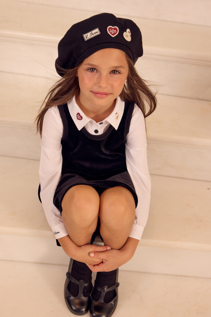 Young girl wearing a black cap, white shirt, and dark dress sitting on a white surface.