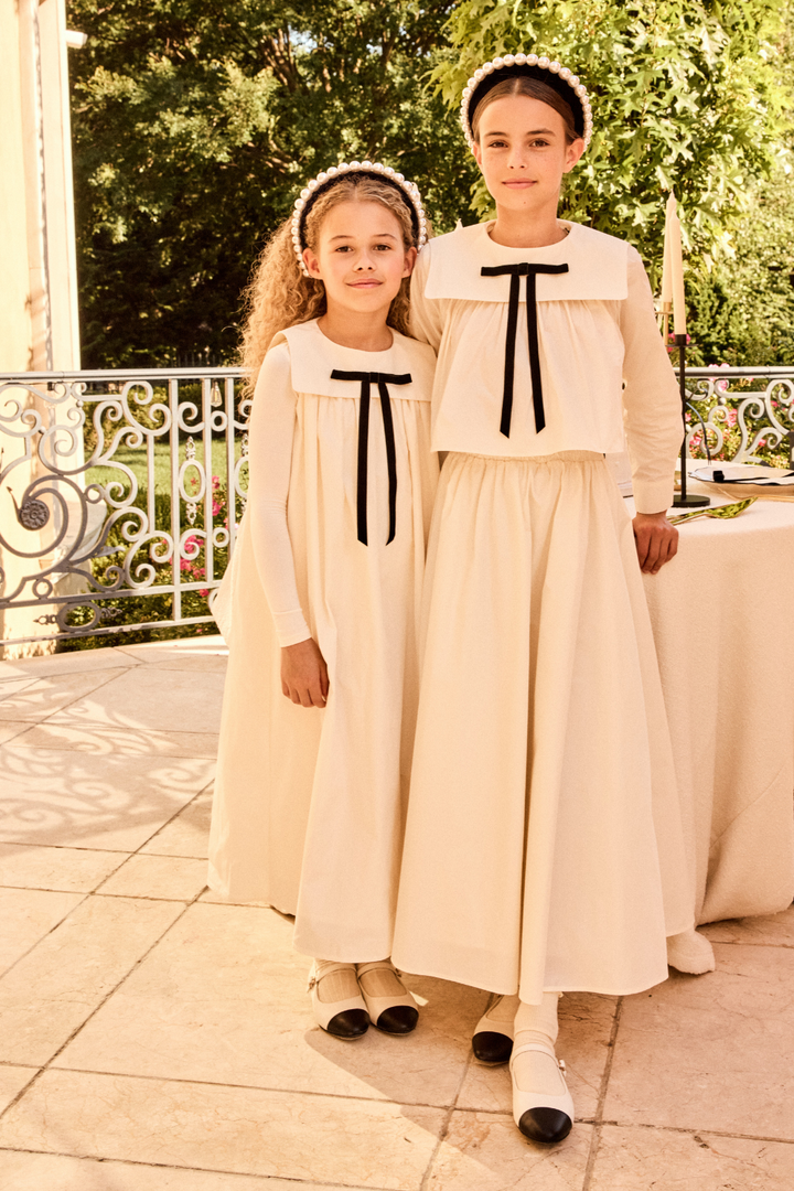 Two children in matching white dresses with black bows standing outdoors.