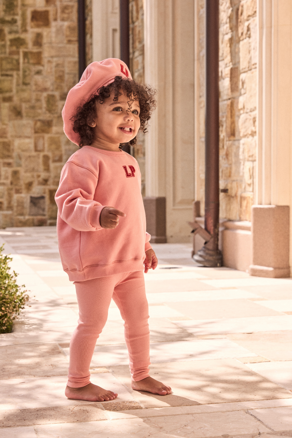Child wearing a pink outfit with a matching hat in front of a stone building.