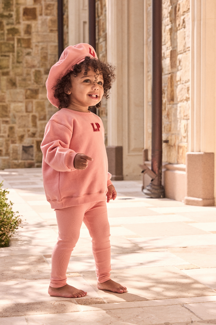 Child wearing a pink outfit with a matching hat in front of a stone building.