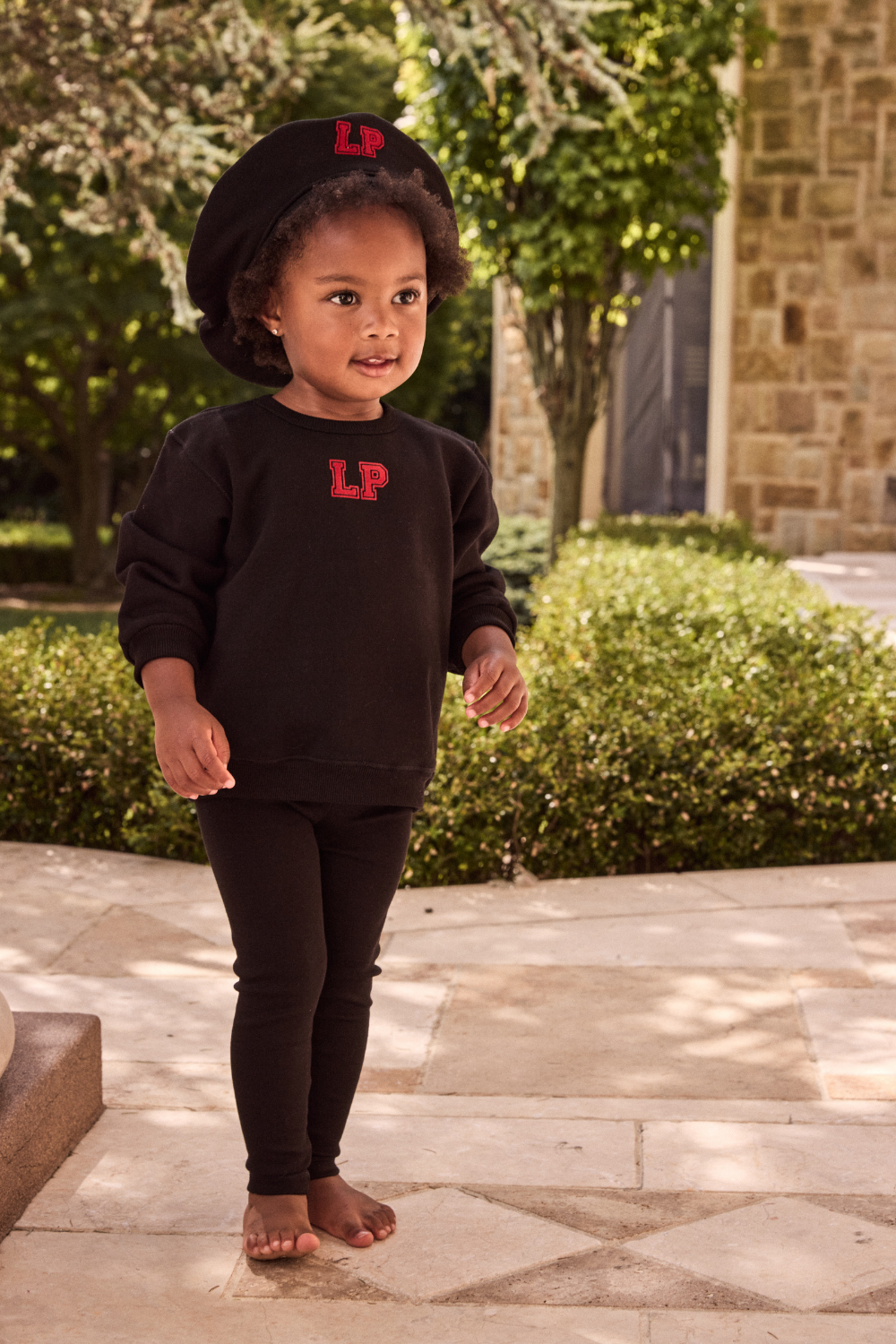 Child wearing a black outfit with 'LP' logo, standing outdoors on a stone path.