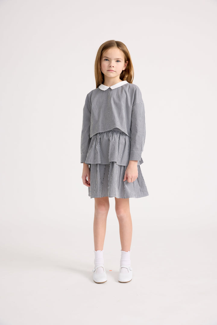 Little girl wearing a cropped long sleeve striped top with a peter pan collar and matching tiered skirt with white socks and flats
