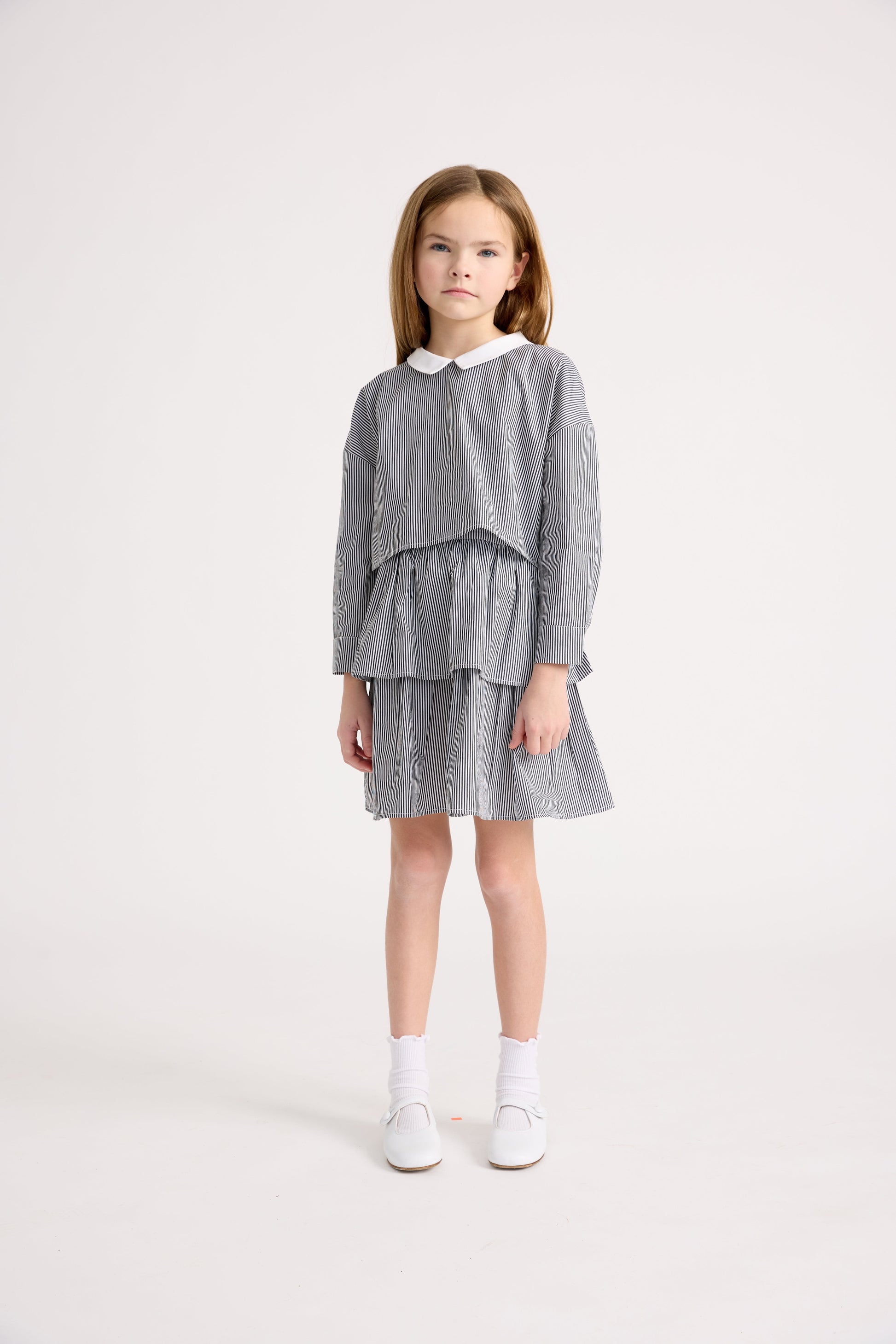 Little girl wearing a cropped long sleeve striped top with a peter pan collar and matching tiered skirt with white socks and flats