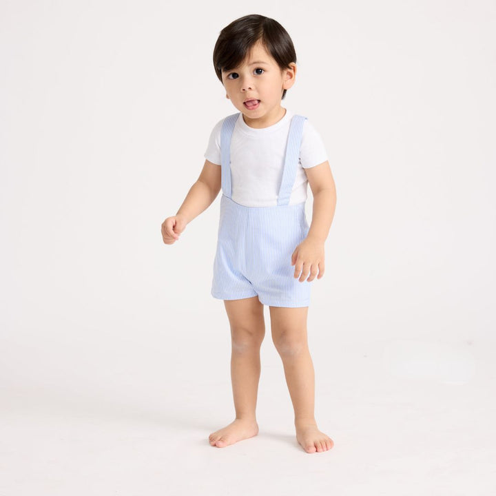 Little boy standing wearing the baby shorts overalls in blue and white stripe with a white short sleeve tee