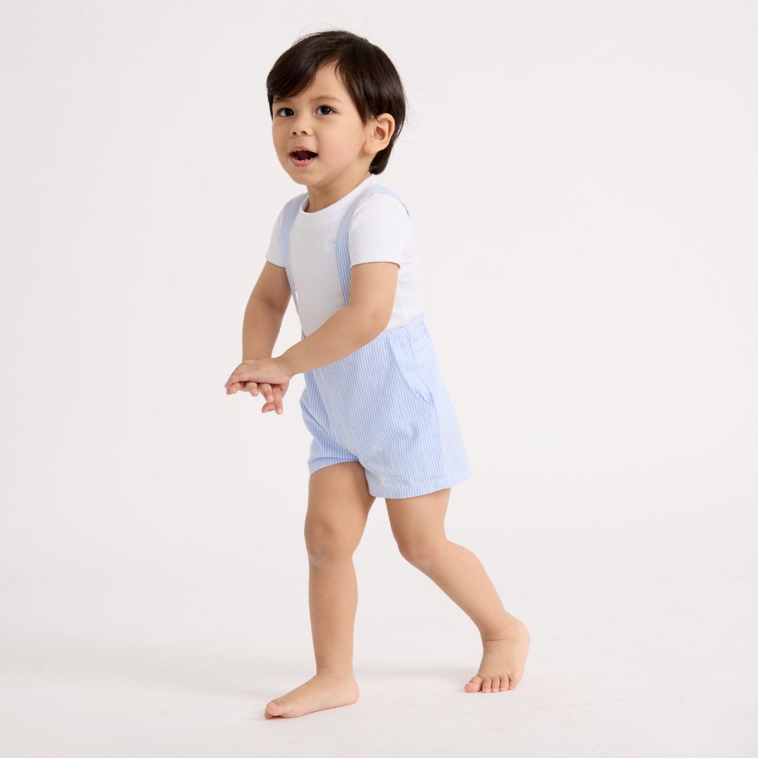 Little boy standing wearing the blue and white striped shorts overalls with a white tee
