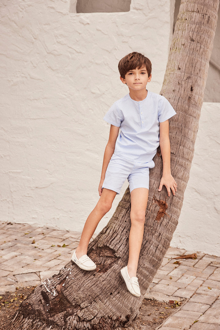 Young boy wearing a striped blue and white collarless shirt with matching shorts leaning against a palm tree