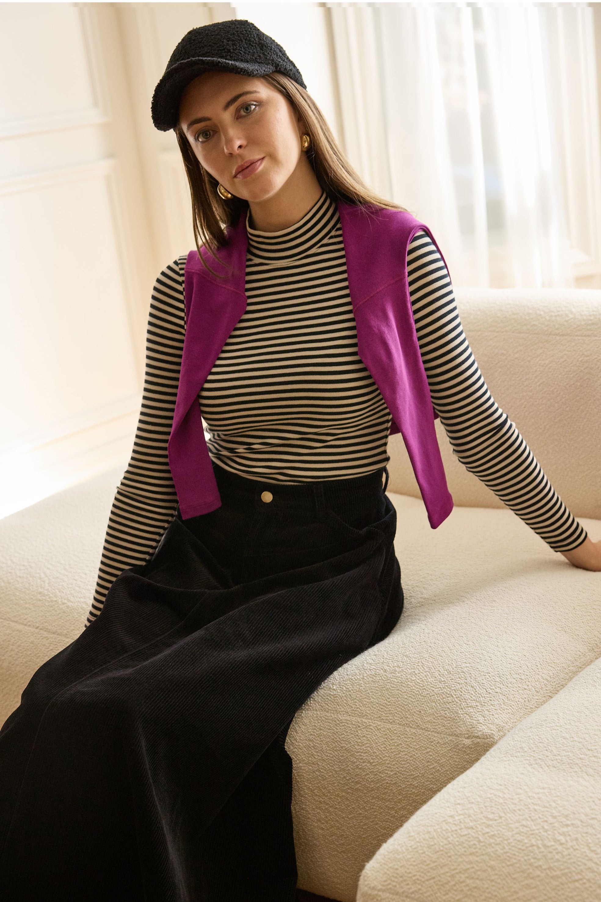 Woman seated wearing striped mockneck, purple sweater on shoulders and black skirt.