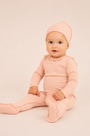 A baby wearing a soft light pink long-sleeve footed one-piece outfit with a matching beanie hat, sitting against a plain light beige studio background.