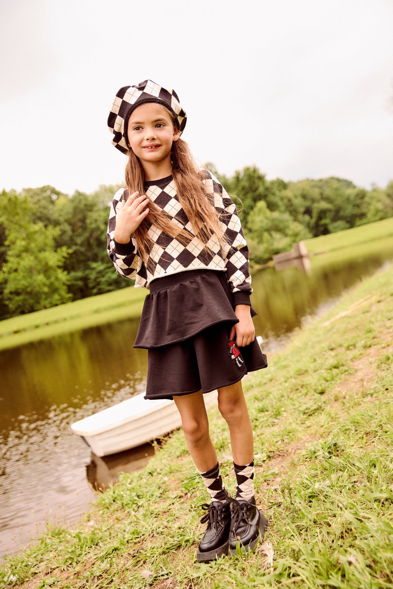 A young girl stands on a grassy bank next to a calm lake, wearing a black and beige argyle sweater, a matching argyle beret, a black skirt, and black lace-up shoes.