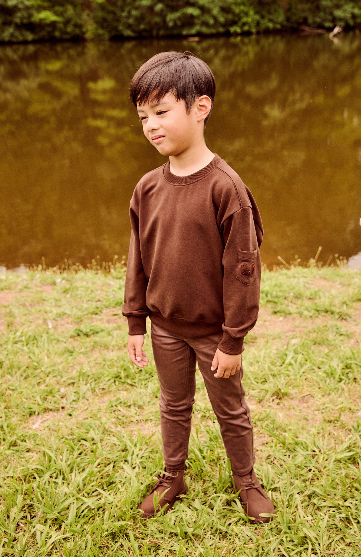 A young boy wearing a matching brown crewneck sweatshirt and jogger pants stands on a grassy lawn with a body of water and trees in the background.