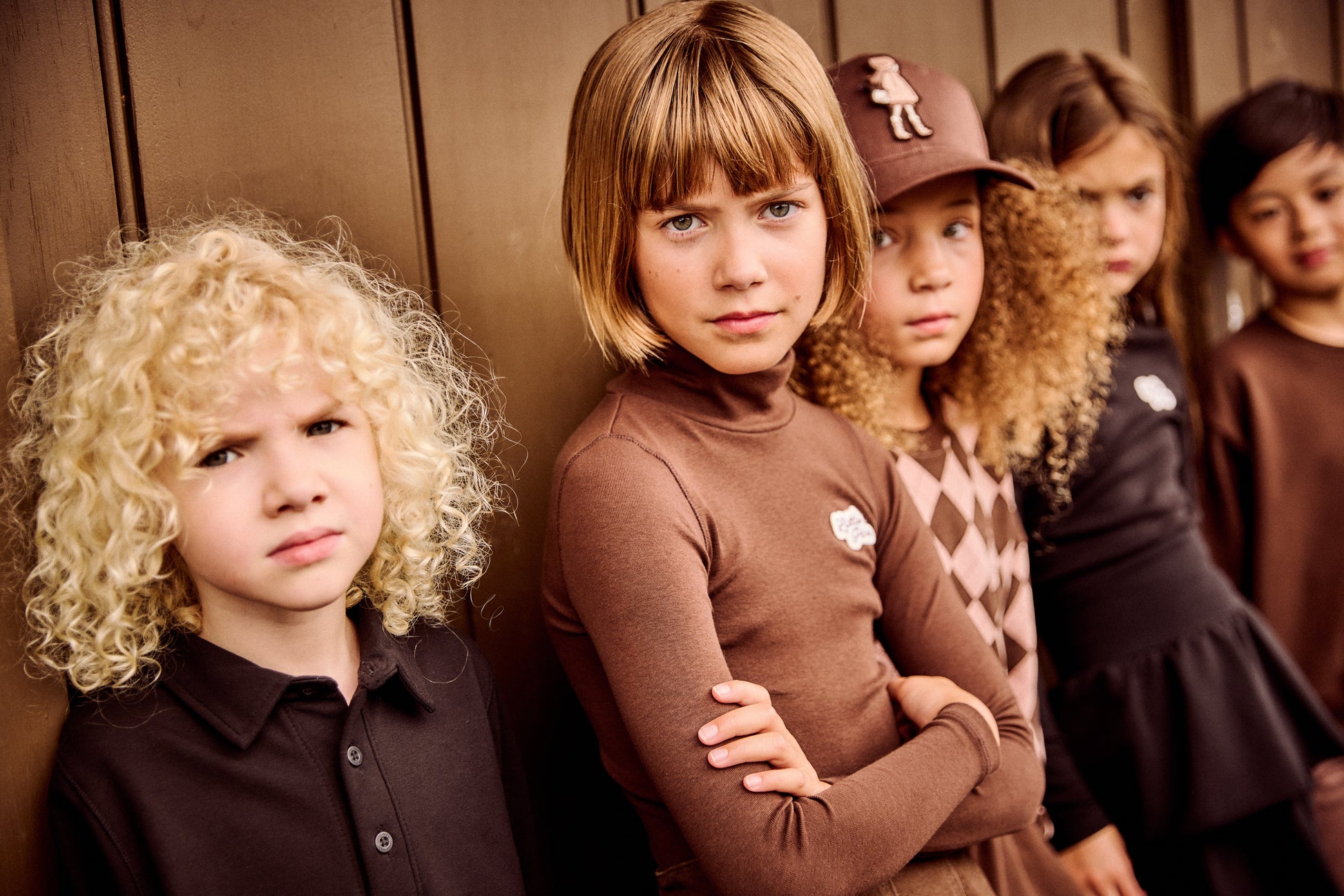 A group of children wearing dark brown and black clothing, standing against a wooden wall. The child in the center wears a brown long-sleeve turtleneck, and another child wears a brown baseball cap with a white graphic.