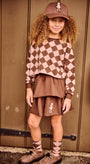 A young girl with curly hair stands against a brown wooden door wearing a matching brown and pink argyle sweatshirt, brown skirt, argyle socks, brown shoes, and a brown baseball cap.