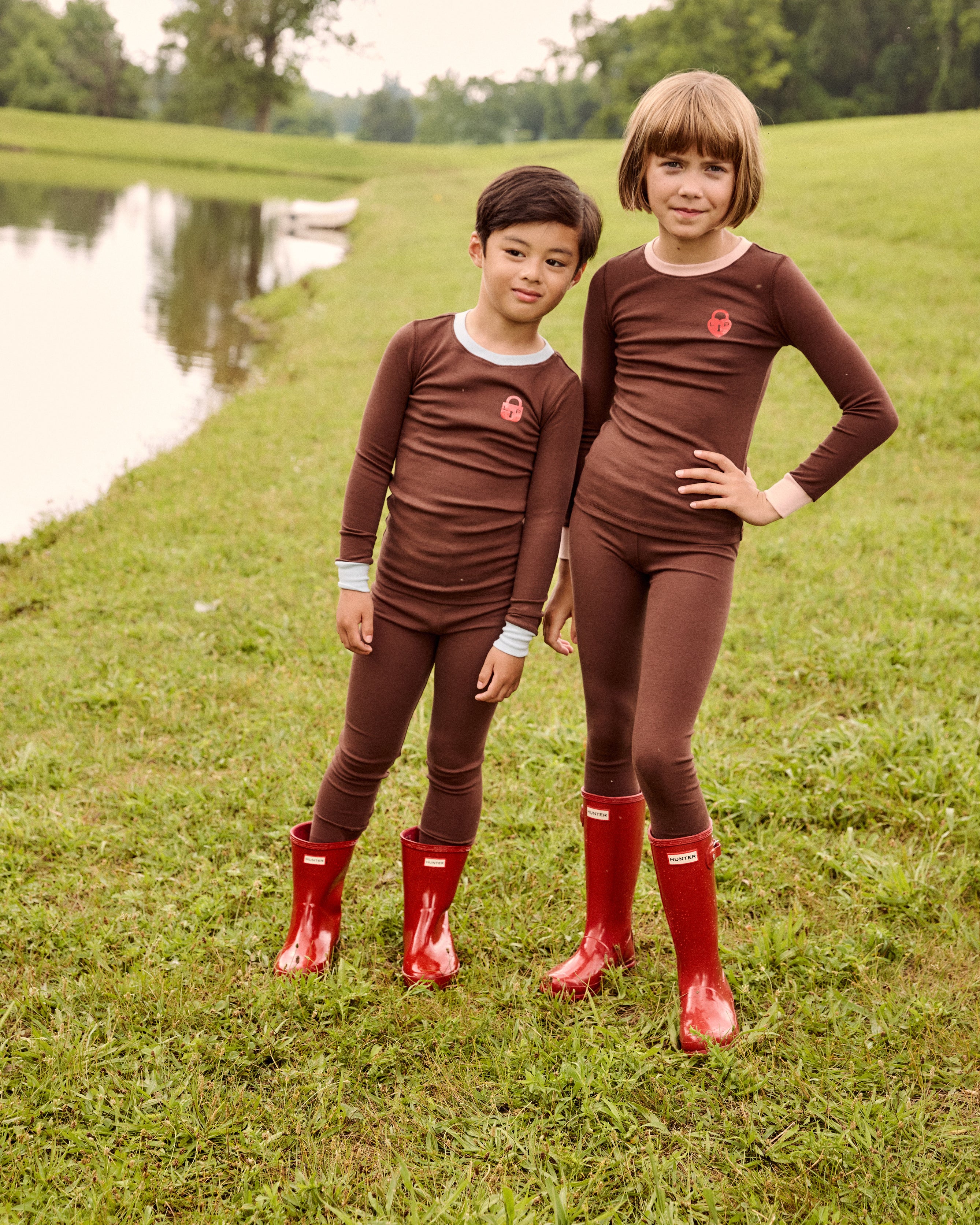 Two children standing outdoors on a grassy field beside a pond, wearing matching brown long-sleeve and matching pajamas, with bright red rain boots. Trees are visible in the background.