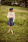 A young girl stands in a grassy field wearing a cream knit sweater with graphics, a blue denim skirt, a blue beret, white socks, and brown shoes.