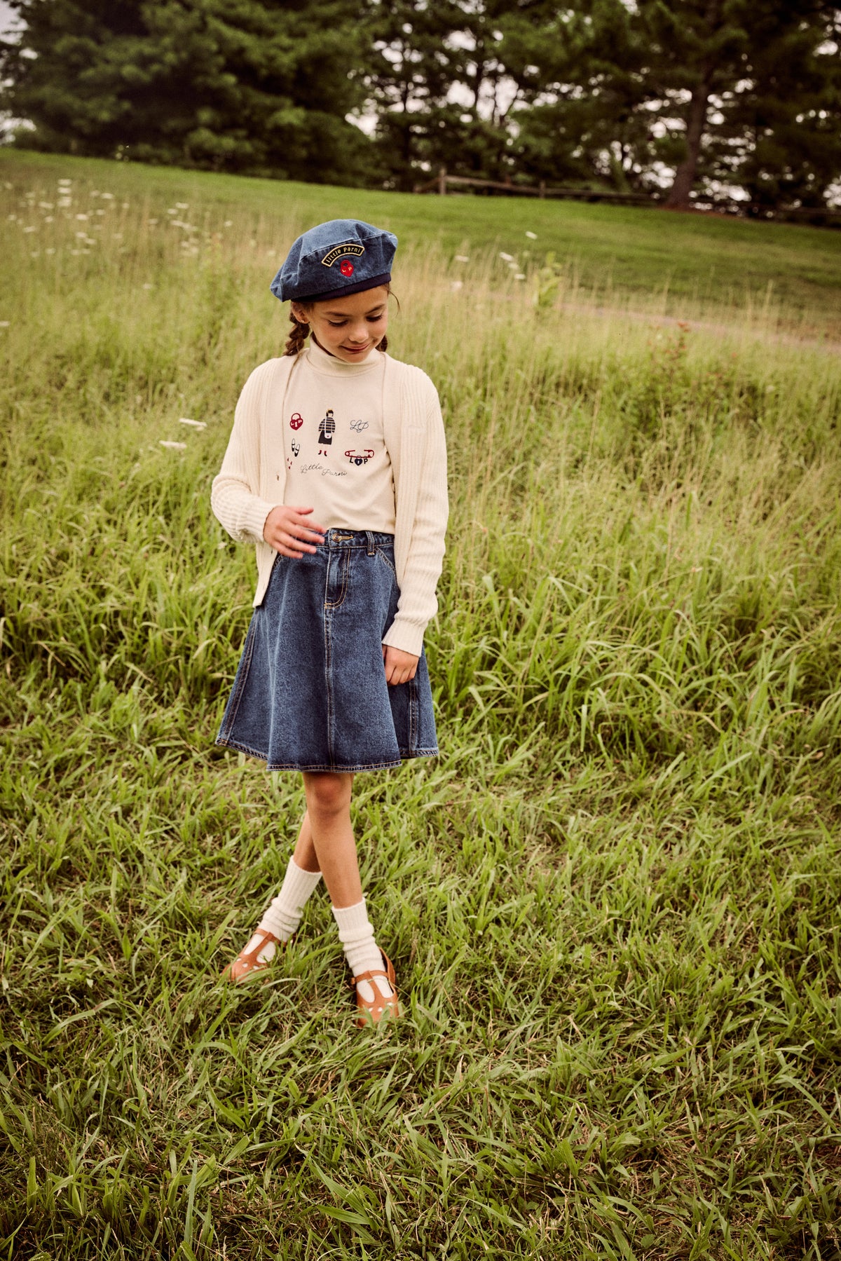 A young girl stands in a grassy field wearing a cream knit sweater with graphics, a blue denim skirt, a blue beret, white socks, and brown shoes.
