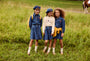 Three young girls standing in a grassy field, wearing matching denim outfits with blue berets, white socks, and sandals. One girl has a yellow jacket tied around her waist, and another has a yellow backpack.