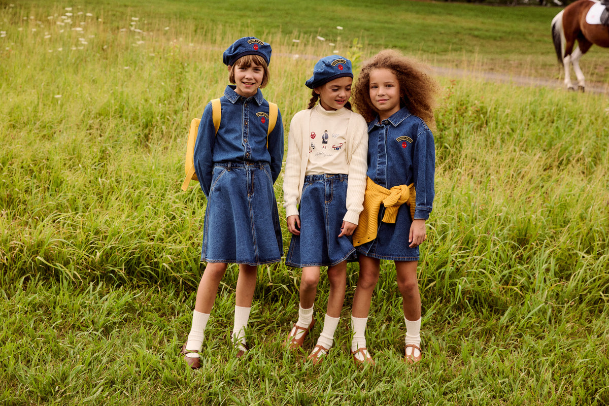 Three young girls standing in a grassy field, wearing matching denim outfits with blue berets, white socks, and sandals. One girl has a yellow jacket tied around her waist, and another has a yellow backpack.