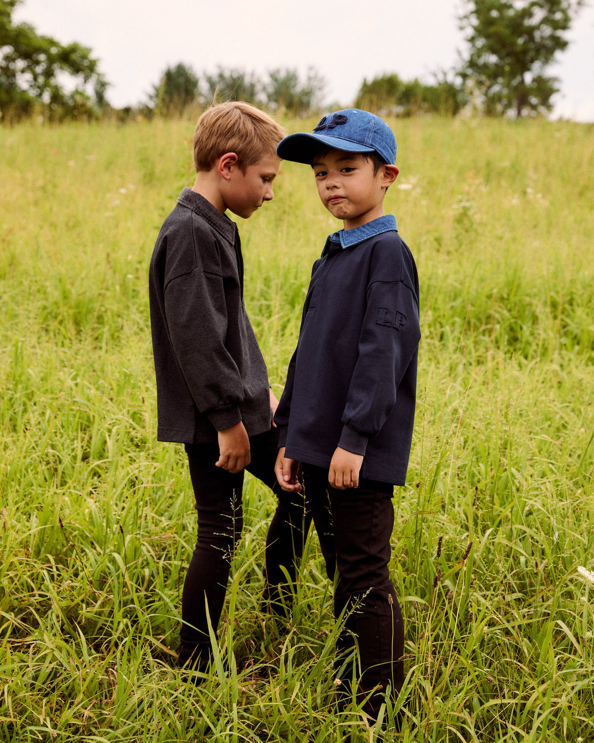 Two young boys standing in a grassy field outdoors. One boy wears a dark gray long-sleeve shirt and dark pants, while the other wears a navy blue long-sleeve shirt, a blue collar, and a blue baseball cap.