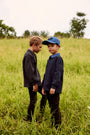 Two young boys standing in a grassy field outdoors. One boy wears a dark gray long-sleeve shirt and dark pants, while the other wears a navy blue long-sleeve shirt, a blue collar, and a blue baseball cap.