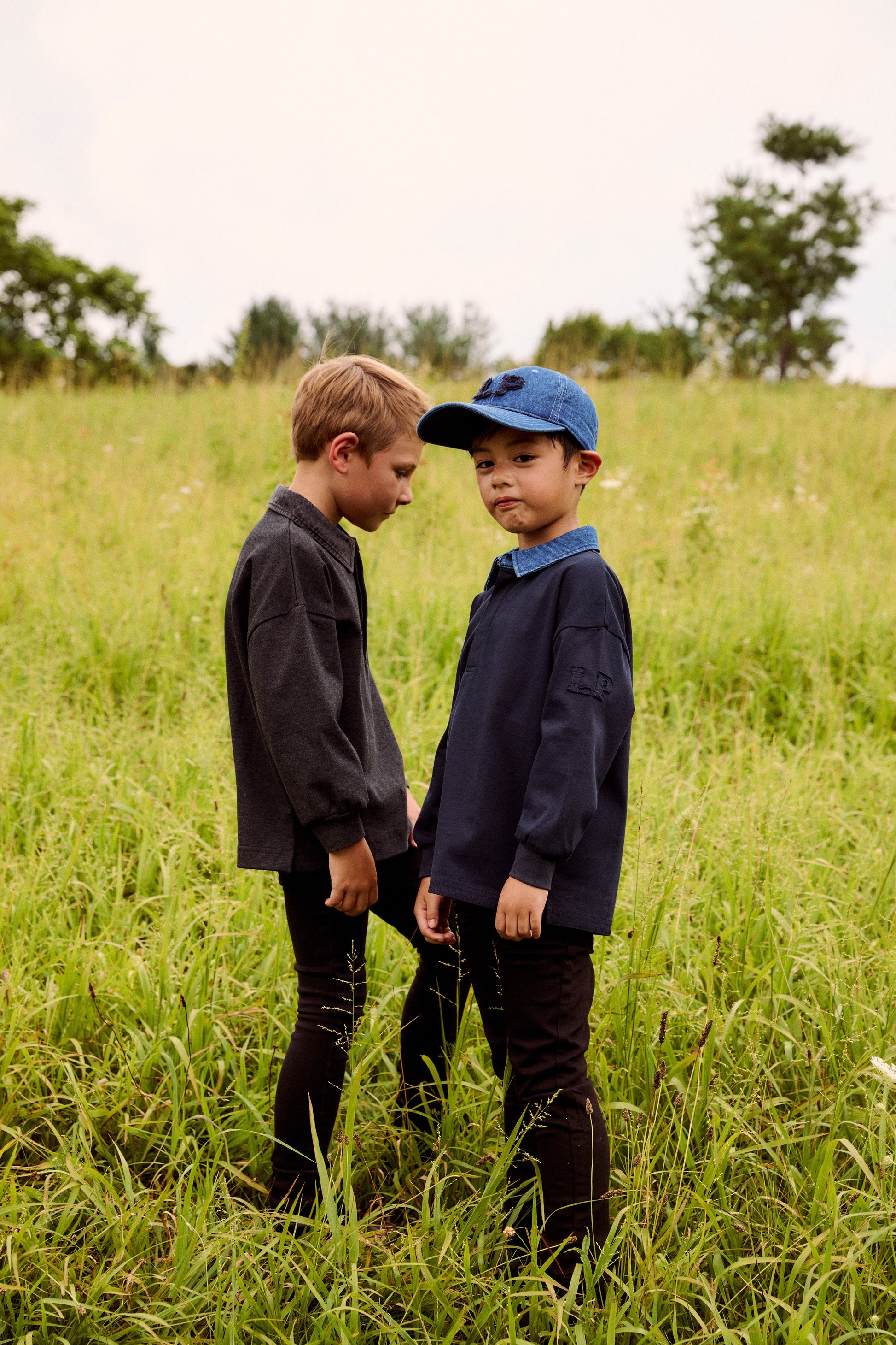 Two young boys standing in a grassy field outdoors. One boy wears a dark gray long-sleeve shirt and dark pants, while the other wears a navy blue long-sleeve shirt, a blue collar, and a blue baseball cap.