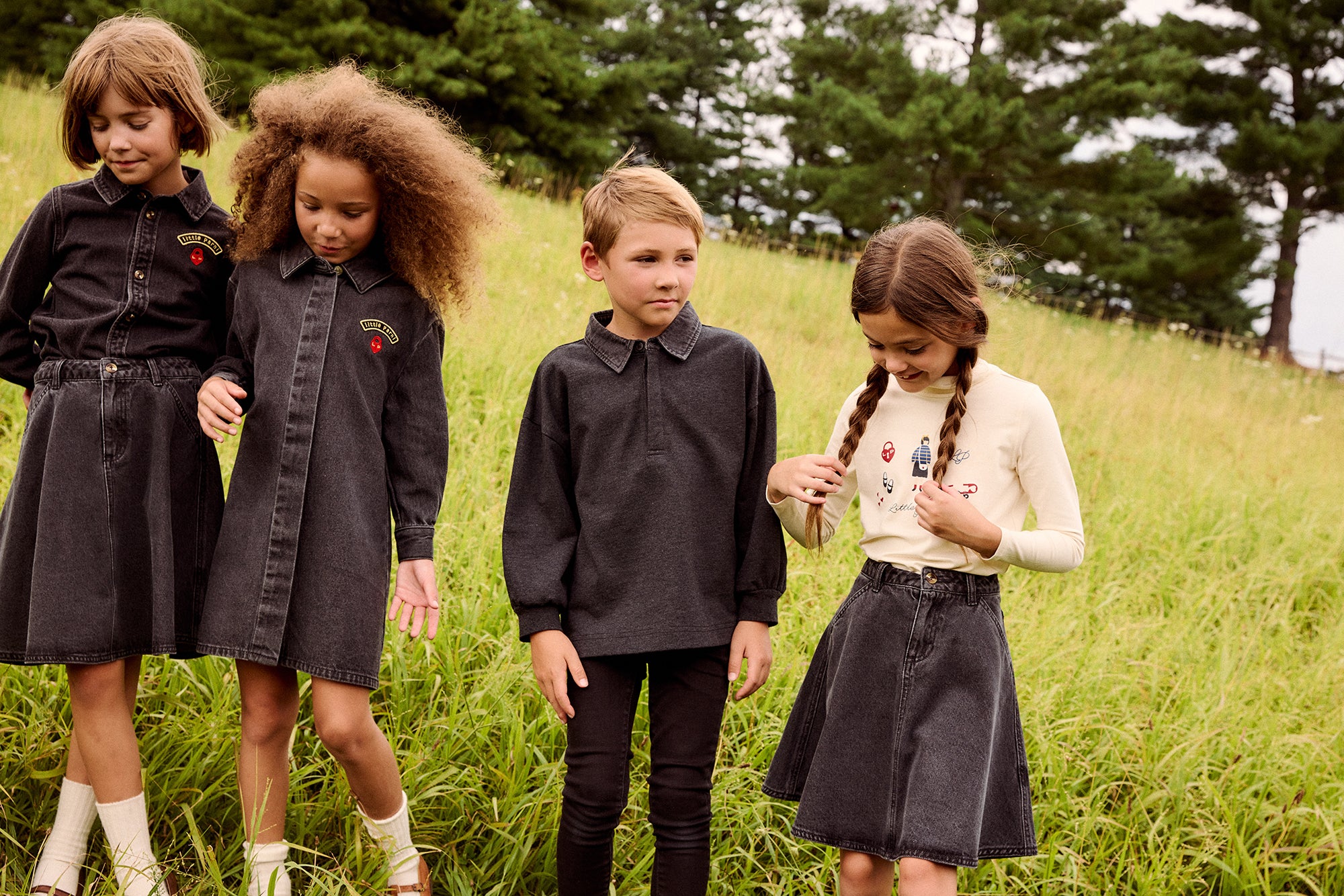 Four children standing in a grassy field outdoors, wearing matching dark gray collared long-sleeve shirts and skirts or trousers. The children are looking down at their clothing, which features small embroidered patches on the chest.