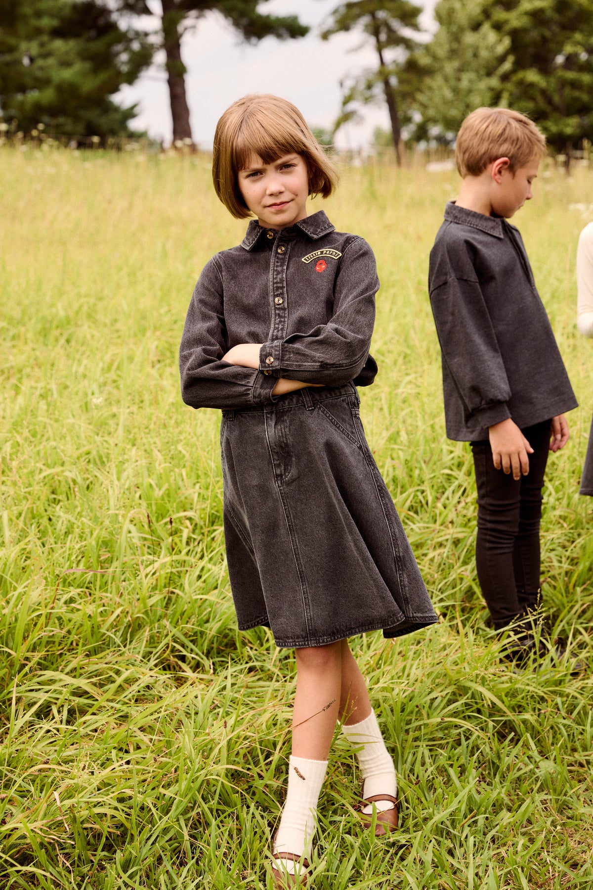 A young girl with a short brown bob stands in a grassy field wearing a dark charcoal gray denim shirt dress, with her arms crossed. A young boy in a matching dark gray shirt and black pants stands in the background.