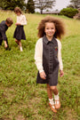 A young girl stands in a grassy field wearing a dark gray denim pinafore dress over a cream knit long-sleeve shirt. Two other children are visible in the background on the hill.