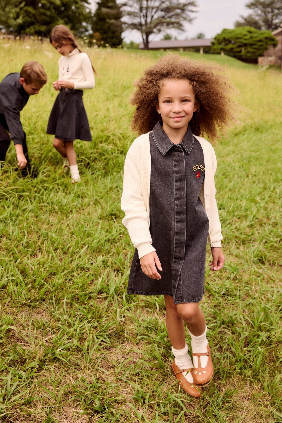 A young girl stands in a grassy field wearing a dark gray denim pinafore dress over a cream knit long-sleeve shirt. Two other children are visible in the background on the hill.